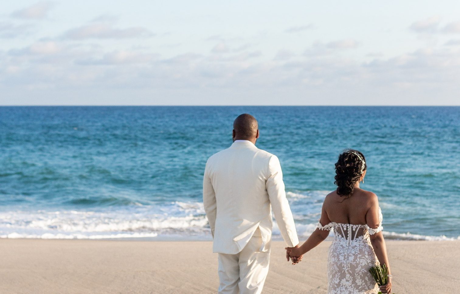 Married couple walks towards ocean in white gown and tuxedo